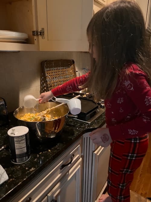A young girl in red pajamas stands on tiptoe at a kitchen counter stirring a large mixing bowl with a measuring cup nearby. She focuses on combining ingredients.