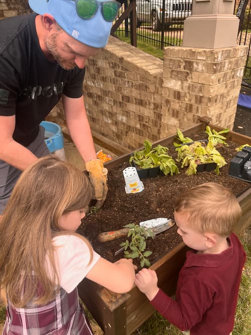 An adult and two children gather around a raised garden bed while planting small seedlings into the soil with a hand trowel.