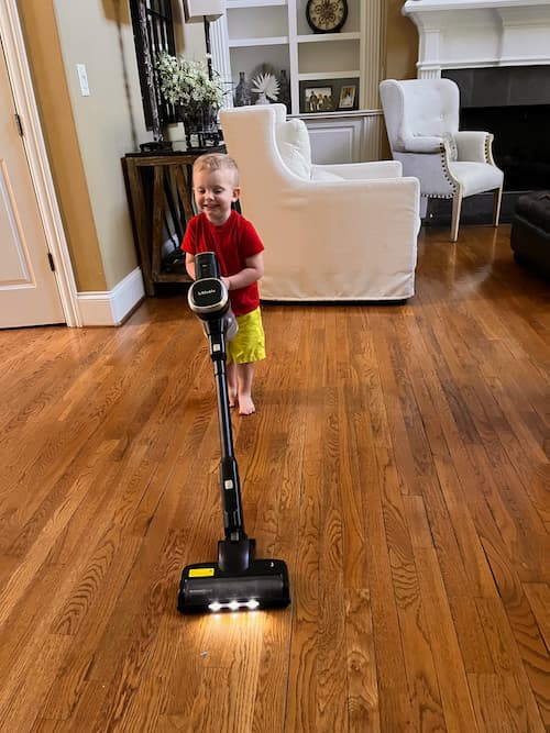 A young boy smiles while pushing a lightweight vacuum across a hardwood living room floor with chairs and a fireplace in the background.