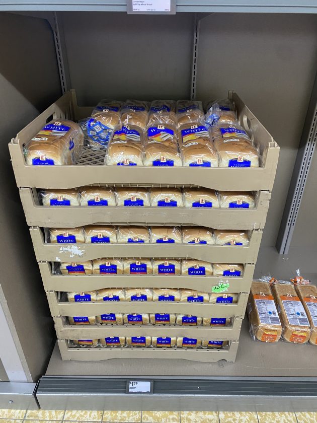 Shelves stocked with packaged white bread loaves arranged in stacked trays.