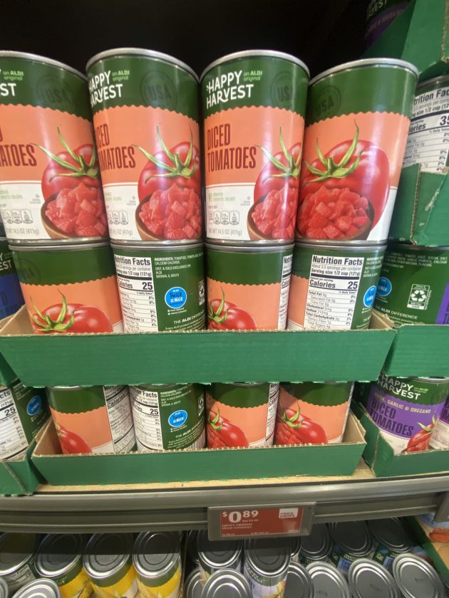 Shelves of canned tomatoes labeled "Happy Harvest Diced Tomatoes" with images of tomatoes on the front and a price sign reading "0.89". These pantry staples are commonly included in best Aldi products lists for budget cooking.