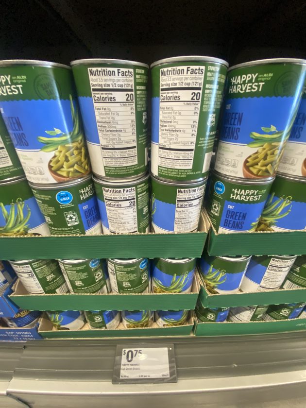 Stacked cans of green beans labeled "Happy Harvest Cut Green Beans" with visible nutrition facts panels and a price tag of "0.75."
