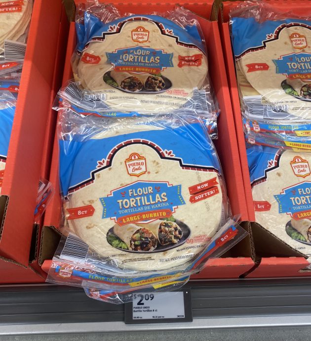 Packs of tortillas labeled "Pueblo Lindo Flour Tortillas Large Burrito 8ct Now Softer" arranged in a display with a price tag below.