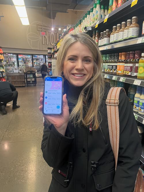 Woman standing in a grocery store aisle smiles while holding up her phone to show an app screen, with shelves of bottled dressings and products behind her.