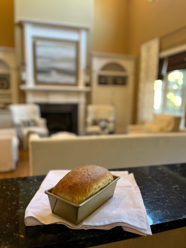 Freshly baked loaf of bread sits in a metal pan on a folded cloth on a kitchen counter.