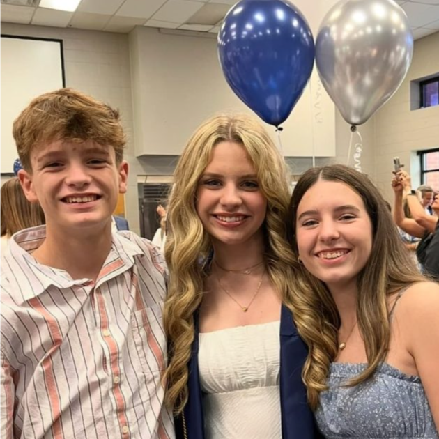 Three teens stand in a room with balloons behind smiling at the camera. One of them is wearing a greaduation gown.