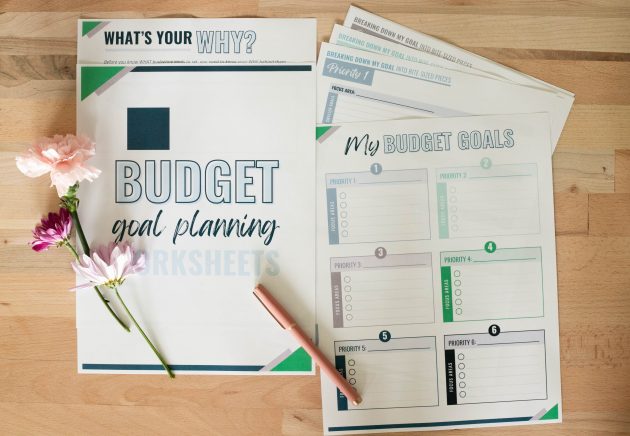 Printed budget goal planning worksheets spread across a wooden table with a pink pen and flowers beside them. The worksheets help organize financial priorities and create a plan for how to get out of debt.