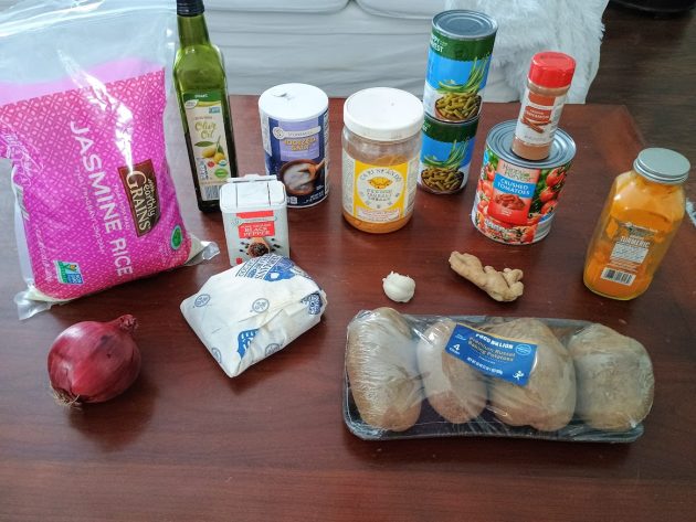 Ingredients laid out on a table including jasmine rice, canned tomatoes, green beans, spices, potatoes, onion, garlic, and ginger for making Pakistani kima.