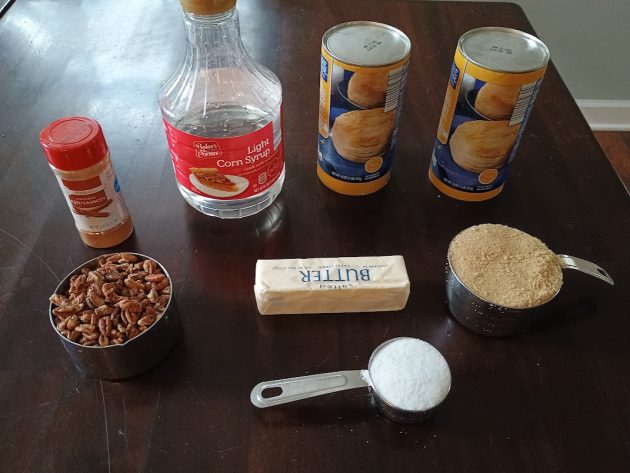 Ingredients for monkey bread recipe including canned biscuit dough, corn syrup, butter, brown sugar, cinnamon sugar, and chopped pecans arranged on a table.