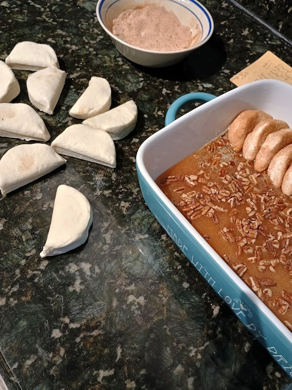 Cut biscuit dough pieces on a countertop next to a baking dish filled with brown sugar and pecans, illustrating how to assemble easy monkey bread before baking.