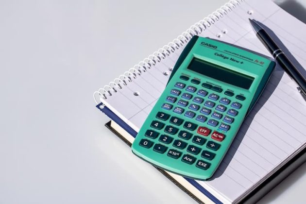 Teal calculator resting on top of a spiral notebook with a pen beside it on a desk.