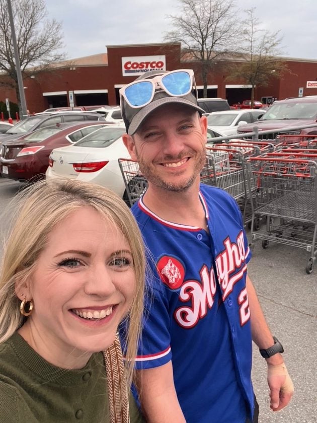 Two smiling people take a selfie in the Costco parking lot with shopping carts and the warehouse entrance behind them.