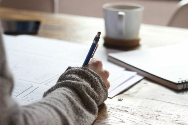 Person writing on paperwork at a wooden table with a coffee mug and notebook nearby.