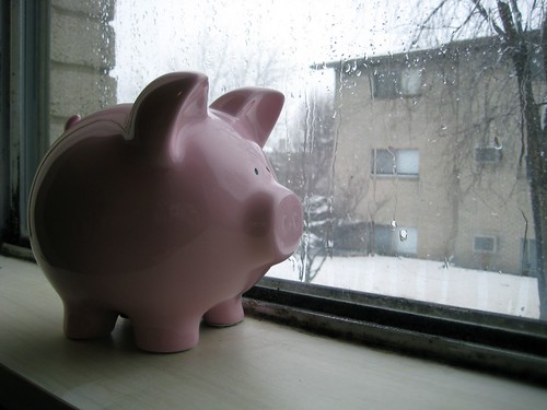 Pink ceramic piggy bank sitting on a windowsill beside a rainy window with buildings visible outside. The classic savings symbol represents making consistent plans for saving money.
