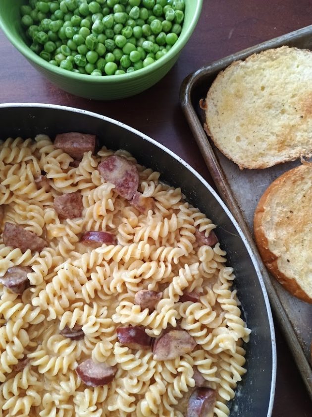 Skillet filled with rotini pasta and sliced kielbasa in a creamy sauce served with a bowl of green peas and toasted bread on the side.