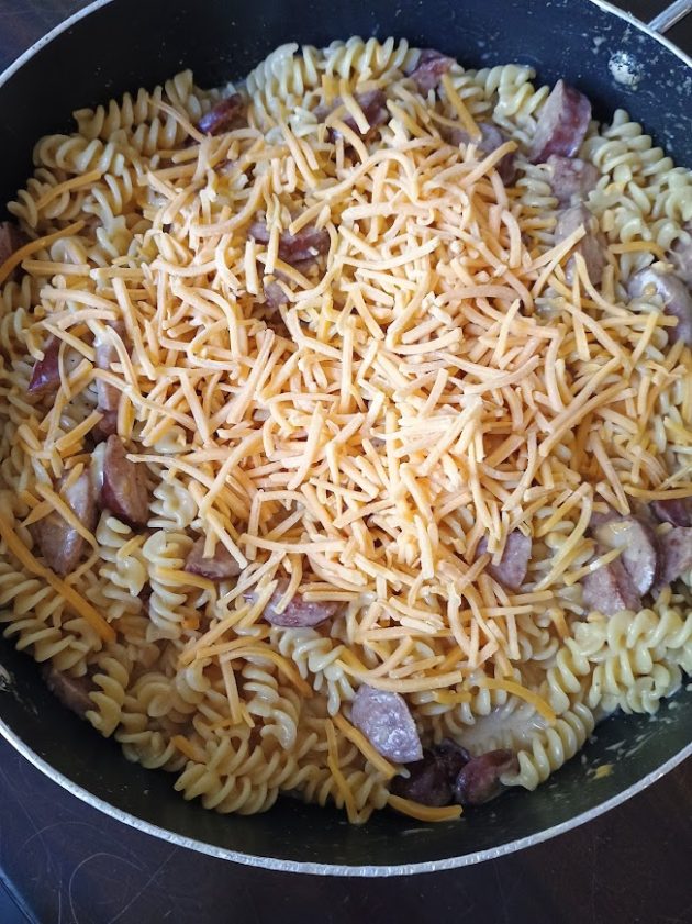 Shredded cheese scattered over hot rotini pasta and kielbasa in a skillet as it begins to melt into the dish for a cheesy kielbasa skillet.