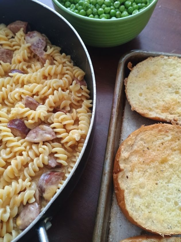 Skillet filled with rotini pasta and sliced kielbasa in a creamy sauce served with a bowl of green peas and toasted bread on the side