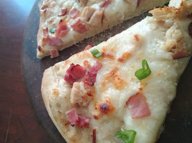 Two slices of chicken alfredo pizza on a wooden board with browned cheese, chunks of chicken, bacon pieces, and green onions.