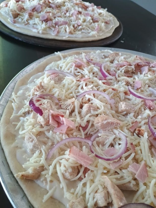 Two uncooked chicken alfredo pizzas on metal trays covered with shredded cheese, chicken, ham, and red onion slices.