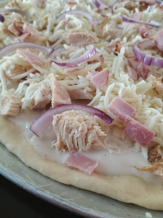 Unbaked chicken alfredo pizza topped with shredded mozzarella, sliced red onions, diced ham, and chunks of chicken over a creamy white sauce.