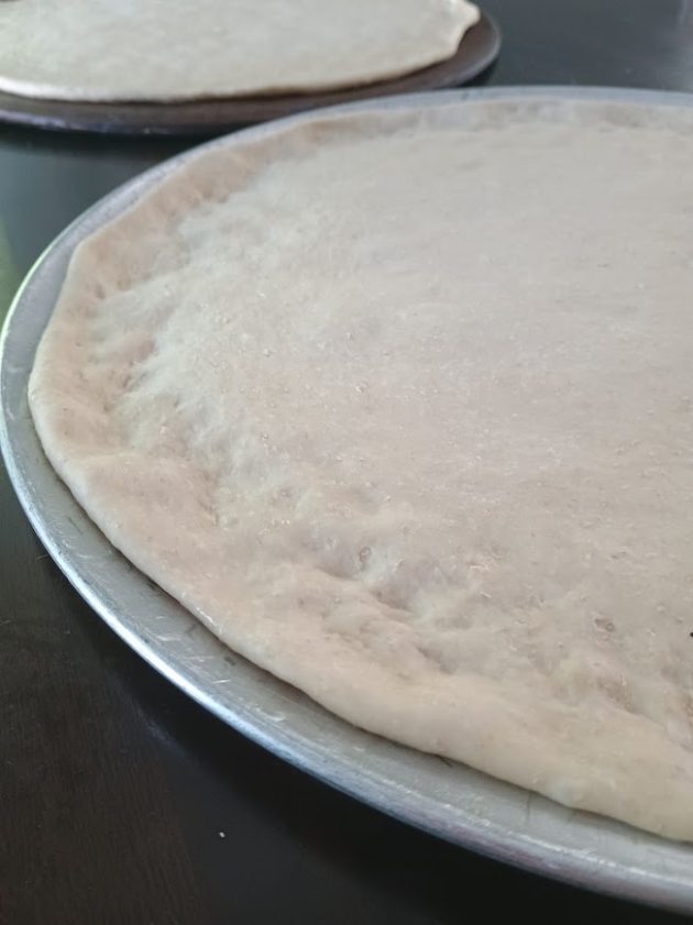Flattened pizza dough spread across a round metal tray with a slightly raised edge forming the crust. The smooth surface is ready for sauce and toppings.