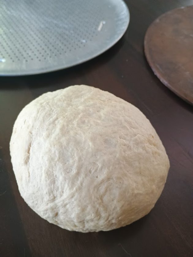 Ball of fresh pizza dough resting on a dark countertop with a soft textured surface. The dough is prepared and ready to be rolled out for a chicken alfredo pizza.
