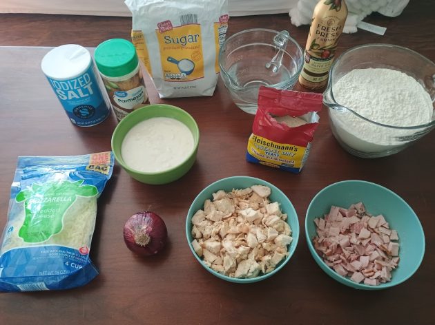 Ingredients for chicken alfredo pizza arranged on a table including shredded mozzarella cheese, diced chicken, chopped ham, red onion, flour, sugar, yeast, olive oil, salt, and Alfredo sauce.
