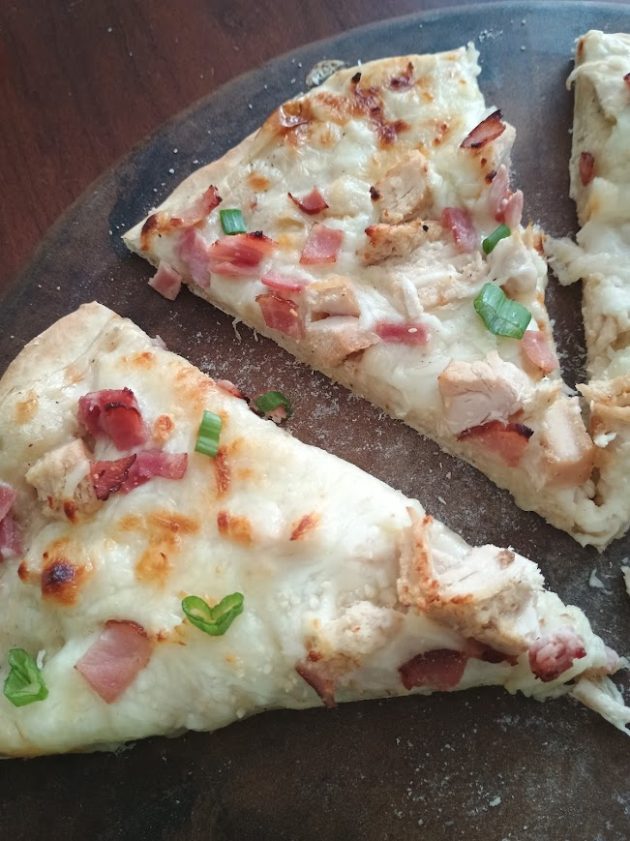 Sliced chicken alfredo pizza arranged on a wooden board with melted cheese, bacon, chicken, and green onions visible on each slice.