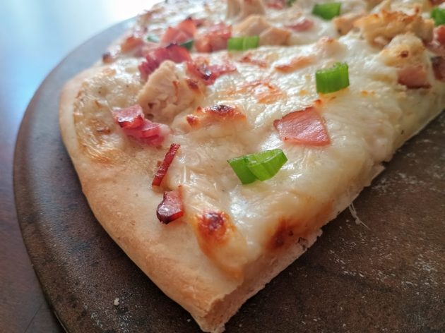 Close up of a slice of chicken alfredo pizza topped with melted mozzarella, diced bacon, grilled chicken, and chopped green onions on a crispy crust.