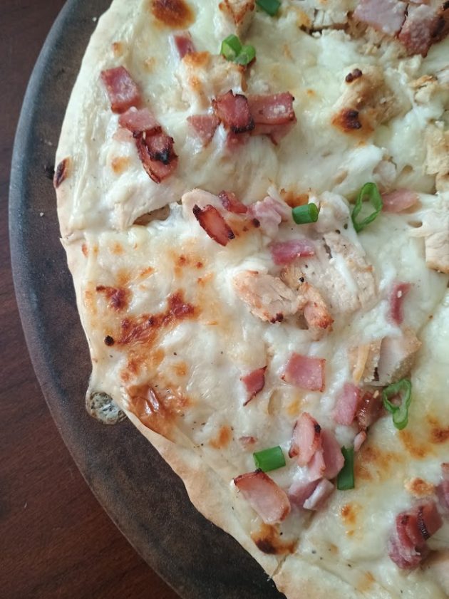 Overhead view of a chicken alfredo pizza with melted cheese, grilled chicken pieces, crispy bacon, and green onion garnish spread across the surface.