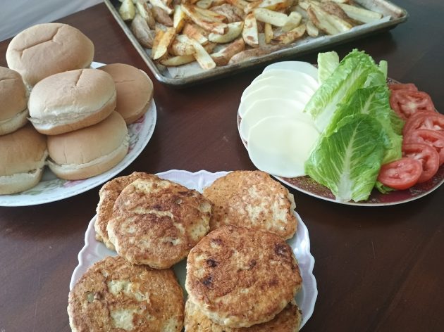 Plate of golden brown chicken patties set out with burger buns sliced cheese, lettuce, tomato, and a tray of fries on a table.