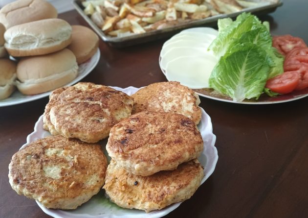 Plate of golden brown chicken patties set out with burger buns sliced cheese, lettuce, tomato, and a tray of fries on a table.
