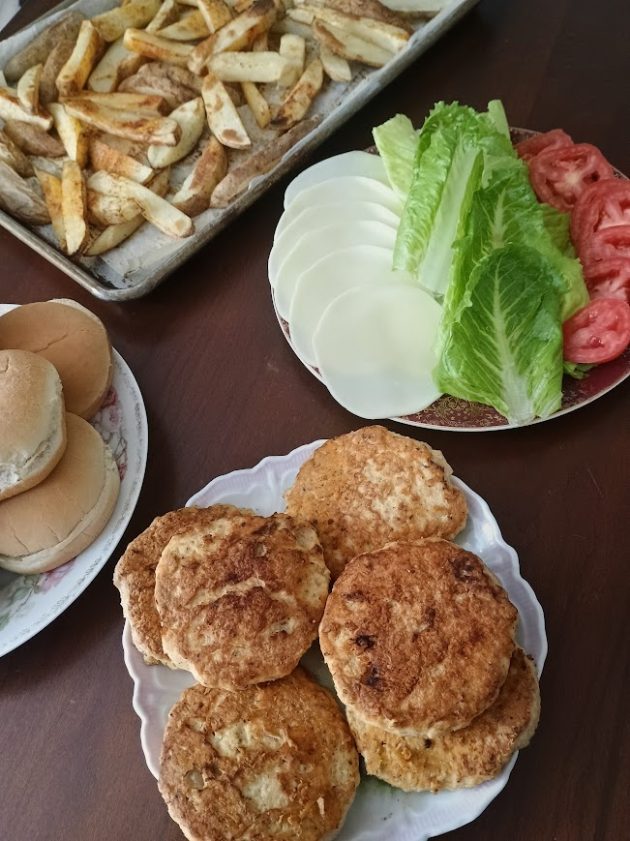 Cooked chicken patties on a plate, beside fresh lettuce, slices of tomato, white cheese, and a tray of fries.