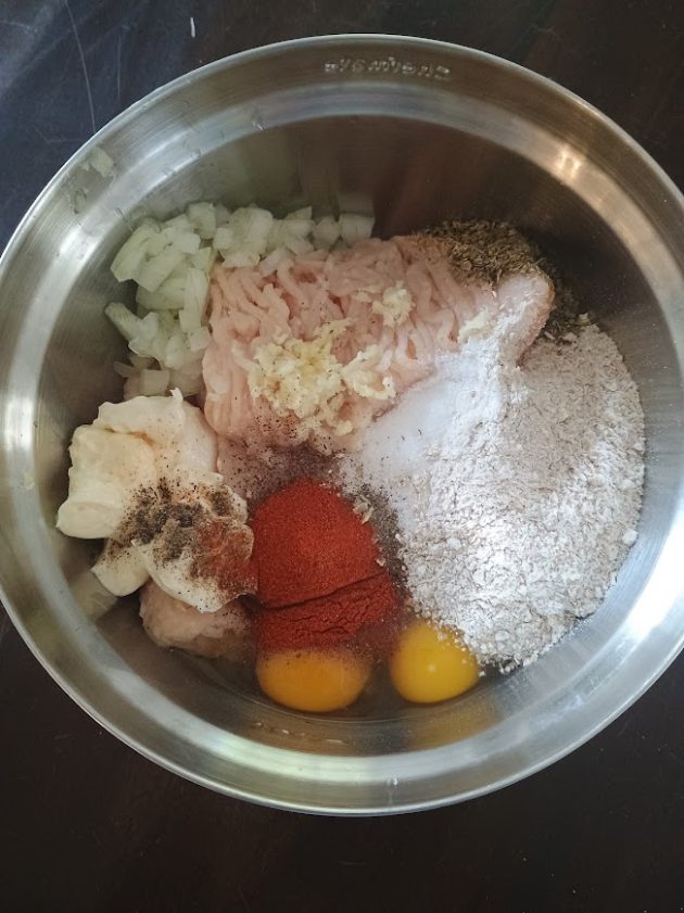 Mixing bowl filled with ground chicken, eggs, flour, chopped onion, garlic, and spices before combining.