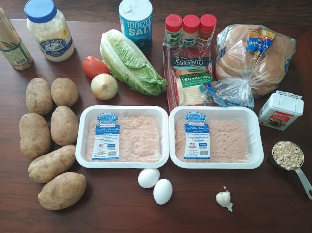 Ingredients laid out including ground chicken, eggs, potatoes, lettuce, tomato, buns, cheese, and seasonings on a table.