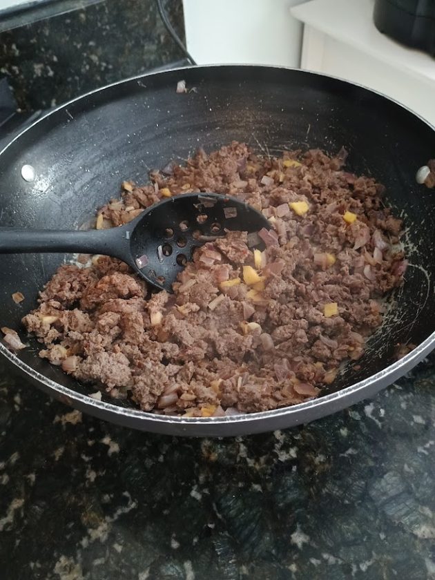 Minced meat being stirred with onions and small pieces of ginger in a pan using a black spoon.
