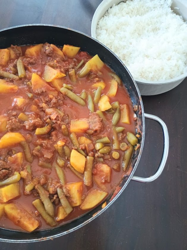 A pan of Pakistani keema curry with chunks of potatoes and green beans in a rich red tomato gravy sits beside a bowl of white rice.