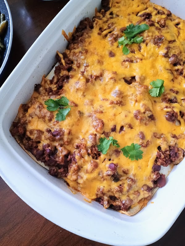 Baked Mexican lasagna topped with melted cheddar cheese and fresh cilantro in a white casserole dish. Layers of tortillas, seasoned meat, and beans make this taco bake a budget friendly family dinner.