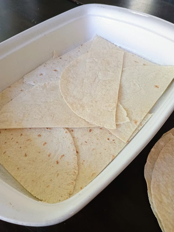 Flour tortillas layered across the bottom of a white casserole dish to prepare homemade Taco Bake.