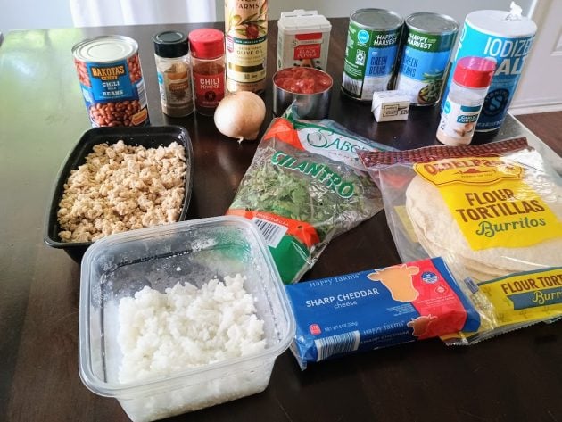 Ingredients for Mexican lasagna arranged on a table including tortillas, shredded cheese, cilantro, canned beans, tomatoes, onions, rice, and seasonings.