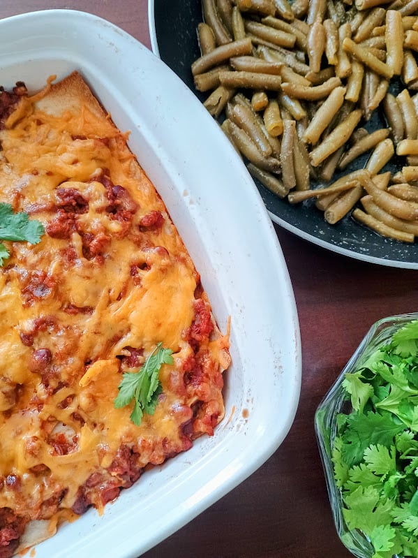 Cheesy taco bake in a white casserole dish served with green beans and fresh cilantro on the side.