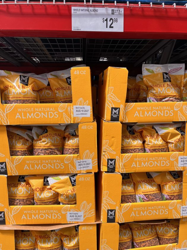 Bulk display of orange boxes labeled "WHOLE NATURAL ALMONDS" with a price sign above reading "$12.98" on a shelf at Sam's Club.