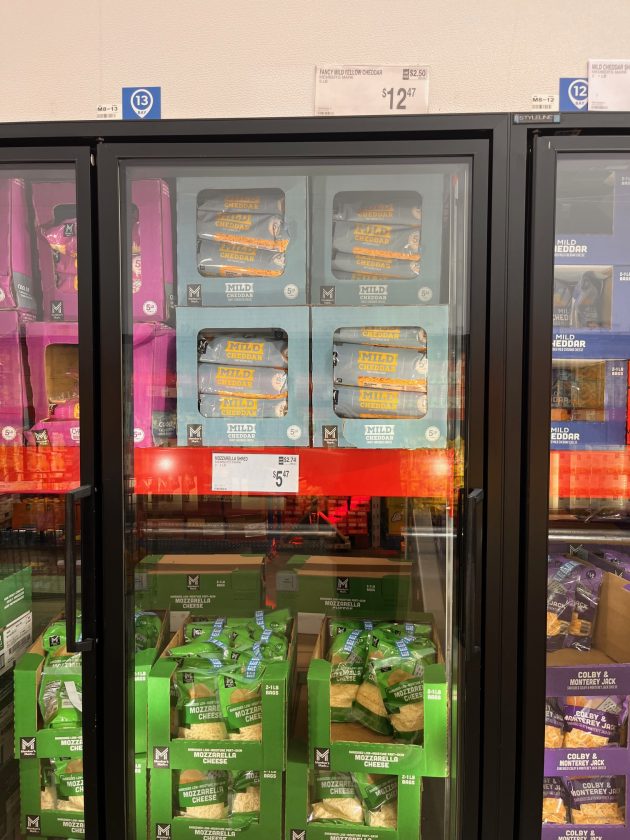 Glass freezer case filled with blocks of mild cheddar and bags of shredded mozzarella cheese stacked in bulk at Sam’s Club with price tags visible above.