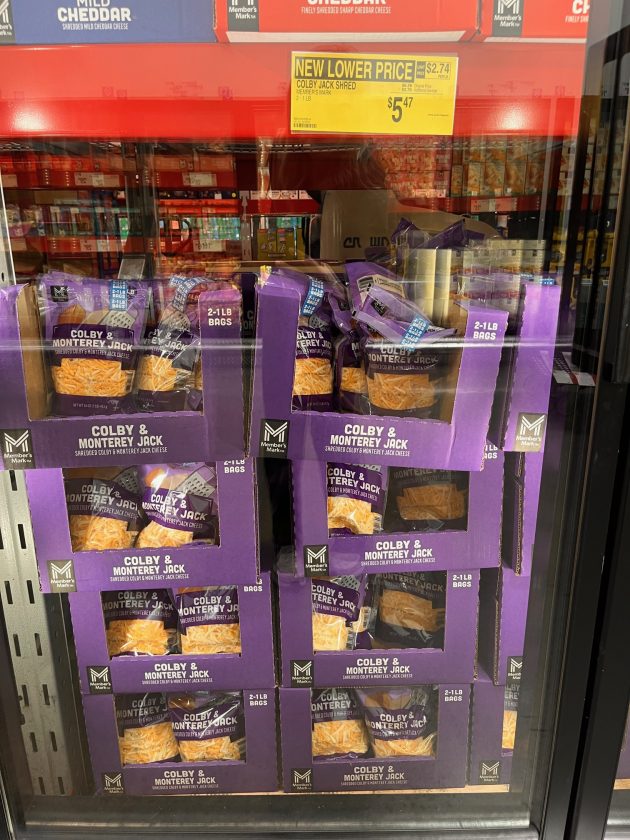 Freezer display of purple bags labeled "COLBY & MONTEREY JACK shredded cheese" with a yellow sign above reading "NEW LOWER PRICE $5.47."