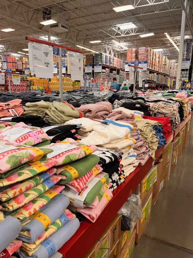 Long table stacked with folded clothing in assorted colors and sizes inside Sam's Club with price signs hanging overhead.