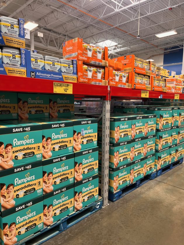Store aisle filled with stacked boxes of Pampers diapers labeled "swaddlers" with signage showing savings above the shelves.