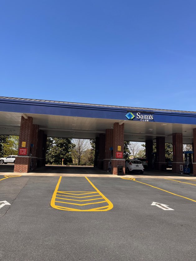 Exterior of Sam’s Club gas station with blue canopy, brick columns, parked cars, and clear blue sky overhead.