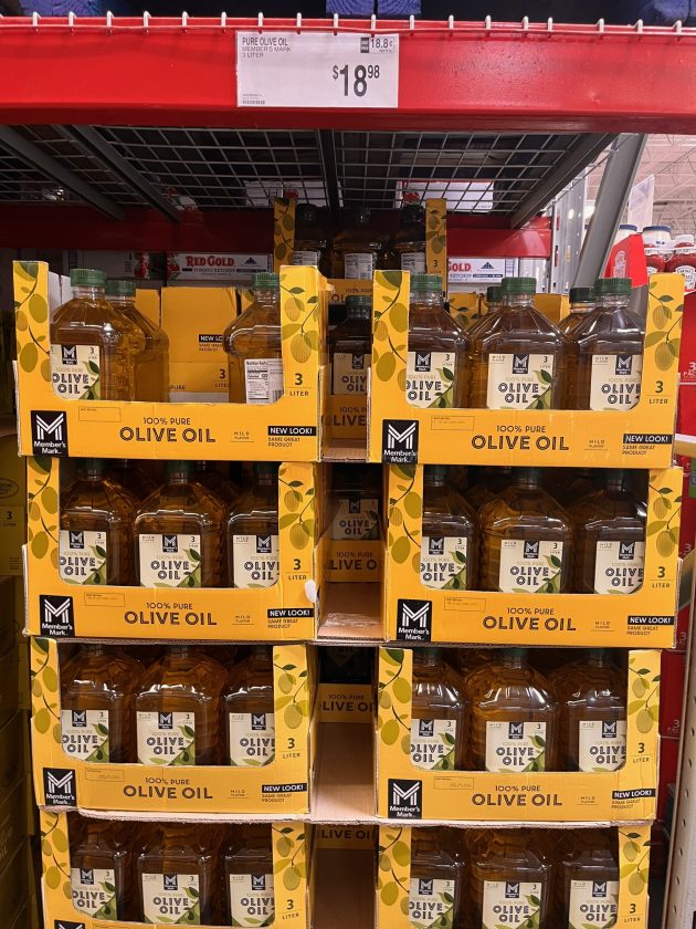 Bulk shelf display of large bottles labeled "100% PURE OLIVE OIL" with a price sign above reading "$18.98."