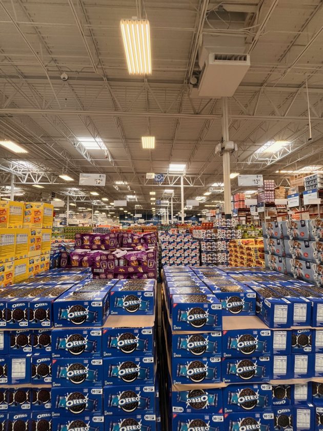 Wide warehouse aisle filled with bulk products including large boxes of "OREO" cookies stacked on pallets under high ceilings.