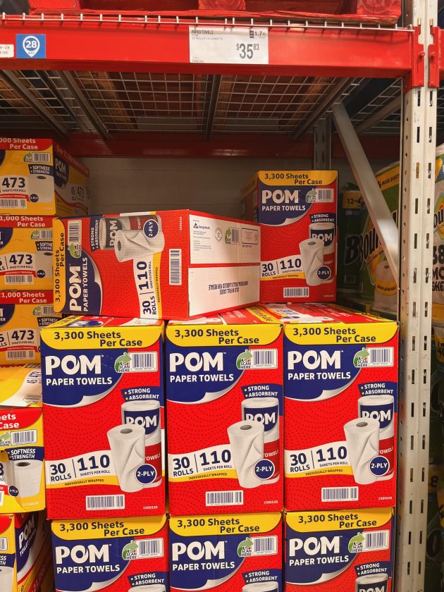 Shelf stocked with boxes labeled "POM paper towels" with text reading "3,300 sheets per case" and "30 rolls 110 sheets per roll.